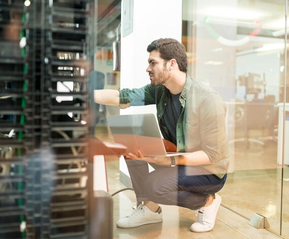 Ein Mann hockt vor einem offenen Serverschrank und hält einen Laptop in einer Hand